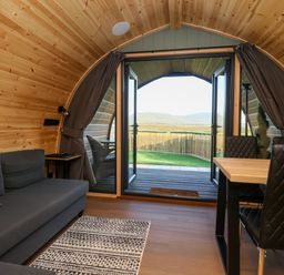 An interior view of a wooden pod with an open double door showing a fenced yard and hills beyond a gray sofa a dining table with black chairs and a rug on wooden floor Curlew Chapel Le Dale near Ingleton