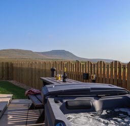A backyard with a bubbling hot tub a picnic table with glasses and a bottle and a wooden fence with hills in the background at Woodpecker in Chapel Le Dale near Ingleton