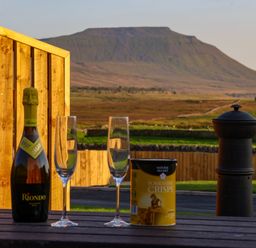 A bottle of sparkling wine two empty glasses and a can of Yorkshire crisps on a wooden table outside with a mountain and green fields in the background at Woodpecker in Chapel Le Dale near Ingleton