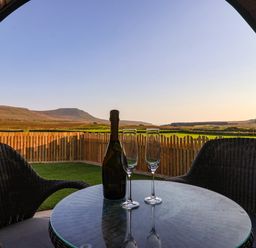 A round glass table with a bottle and two empty champagne glasses between two wicker chairs overlooking a fenced grassy area and distant hills at Woodpecker in Chapel Le Dale near Ingleton