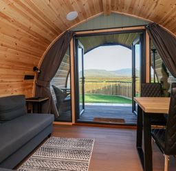A wooden interior room with a gray sofa a table with black chairs and an open door showing a fenced yard and hills at Woodpecker in Chapel Le Dale near Ingleton