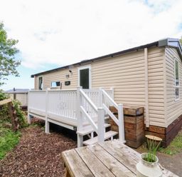 A beige mobile home with a white railing veranda and steps next to a wooden table with a plant pot at 25 Manleigh Park in Combe Martin