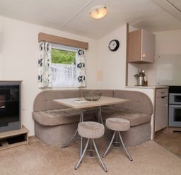 A small dining area with a corner bench sofa table and two stools next to a kitchen with an oven and a wall clock at 26 Manleigh Park in Combe Martin