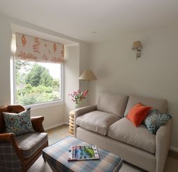 A living room with a beige sofa and an armchair next to a window with floral curtains at Little Robin Cottage in Ambleside