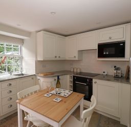A kitchen with white cabinets a wooden table with two chairs and a window with flowers at Little Robin Cottage in Ambleside