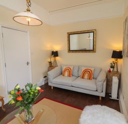 A living room with a grey sofa two orange pillows a mirror and flowers on a glass table at Belmont House in Bowness-On-Windermere