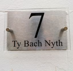 A metal house number sign with the number 7 and the text Ty Bach Nyth on a white wall at Hole in the Wall in Caernarfon