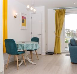 A dining area with two teal chairs and a round table with a patterned tablecloth next to a sliding glass door with yellow curtains and a blue sofa at Rolles Rainbow in Exmouth