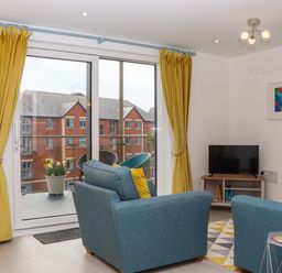 A living room with blue armchairs yellow curtains a small TV and a balcony at Rolles Rainbow in Exmouth