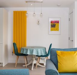 A dining area with a round table covered with a patterned cloth and two teal chairs a yellow vertical radiator on the wall a blue armchair with a yellow cushion and a framed artwork on the wall at Rolles Rainbow in Exmouth