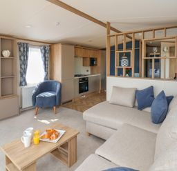 A living room with a beige sofa blue armchair wooden coffee table with croissants and drinks and an open kitchen at Aspen Lodge in Whitby