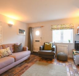 A living room with a pink sofa a gray armchair a TV a brick floor and a fireplace at Caines Cottage in Westleton