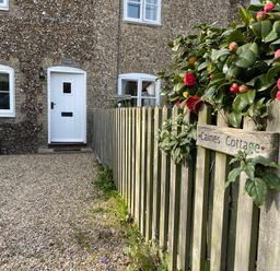 A stone cottage with white door and windows next to a wooden fence with flowers at Caines Cottage in Westleton