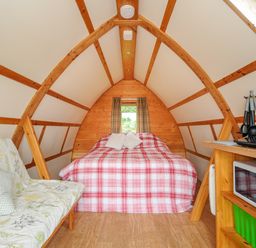 A bedroom with a bed, small window, sofa with cushions, microwave, and kitchen utensils in a wooden cabin at Badger Set @ Penbugle Organic Farm St Keyne near Duloe