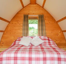 A bed with red and white plaid bedding and two white pillows inside a wooden cabin with a window and beige curtains at Badger Set @ Penbugle Organic Farm in St Keyne near Duloe