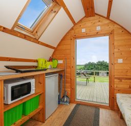A small wooden room with a kitchenette including a microwave fridge and stove and an open door leading to a deck with picnic bench at Badger Set @ Penbugle Organic Farm St Keyne near Duloe