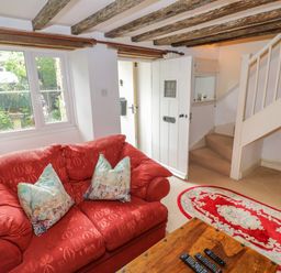A living room with a red sofa two pillows wooden coffee table with remotes oval rug stairs and a white door at 4 Lansdown Cottages Bourton-On-The-Water