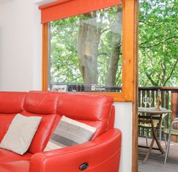 A red sofa with pillows near a window and outdoor balcony with table chairs and bottle at Lodge Nine in Finlake Holiday Park near Chudleigh