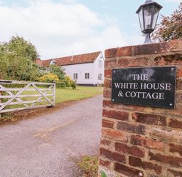 A brick pillar with a sign saying The White House and Cottage next to a white wooden gate and a driveway leading to a white house