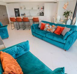 An open living room and kitchen with teal sofas orange cushions kitchen island with orange bar stools and a glass dining table at 17 Cliff Edge in Newquay