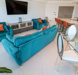 A living room with teal sofas orange cushions a glass dining table with white chairs and an open kitchen with orange bar stools at 17 Cliff Edge in Newquay