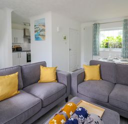 A living room with two gray sofas with yellow pillows a small kitchen and a dining table by a window at Sea Rose in Aberporth
