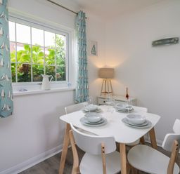 A dining area with a white table set for four and light blue curtains with seagull prints at Sea Rose in Aberporth