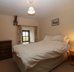 A bedroom with a double bed between a wooden chest of drawers and a wooden nightstand with a lamp and a small window with curtains at Ereiniog 2 in Tremadog