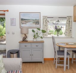 A dining area with a round wooden table and chairs a sideboard with a plant and lamp and a window with floral curtains at Dove Cottage Queens Nympton near South Molton