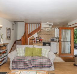 A living room with a sofa and pillows in front of a staircase next to a kitchen and wooden door at Dove Cottage in Queens Nympton near South Molton