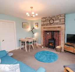 A living room with a blue sofa a fireplace a dining table and chairs and a television on a wooden stand at Woodcutters Cottage Park Village near Haltwhistle