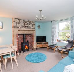 A living room with a stone fireplace wood stove blue round rug dining table chairs television and a window at Woodcutters Cottage in Park Village near Haltwhistle