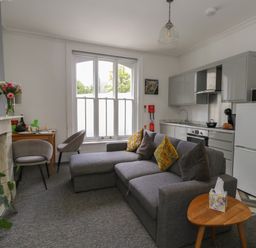 A living area with a gray sofa, two chairs, a small table with tissues, a kitchen with white cabinets, a refrigerator, microwave, and a window with white blinds at Flat 2 - Studland in Bournemouth