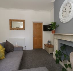 A living room with a gray sofa yellow and gray cushions a wooden door a wall clock plants and a marble fireplace at Flat 2 - Studland in Bournemouth