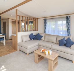 A living room with a beige sectional sofa and blue cushions next to a wooden coffee table with drinks a dining area with a table and chairs and a blue accent wall at Willow Lodge in Whitby