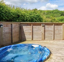 A wooden deck with a blue hot tub and wooden fencing surrounded by green trees at 1 Hedgerows in Lanreath