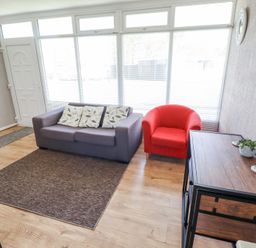 A living room with a gray sofa with three pillows a red armchair a wooden cabinet with a plant and coasters on top and large windows at 68 Cherry Park in Chapel St Leonards