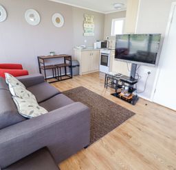 A living room with a gray sofa pillows a red chair tv on a stand a small dining table and kitchenette at 68 Cherry Park in Chapel St Leonards