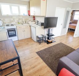 A small kitchen area with stove and refrigerator next to a living room with a couch and TV at 68 Cherry Park in Chapel St Leonards
