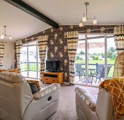 A living room with leather sofas a TV on a wooden stand striped curtains and a garden view at Owls Retreat Lodge 11 Near Aldeburgh
