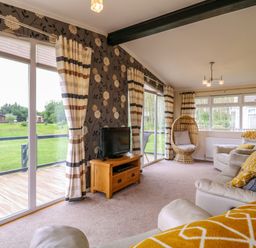 A living room with beige sofas and a wooden TV stand near large windows with striped curtains at Owls Retreat Lodge 11 Near Aldeburgh