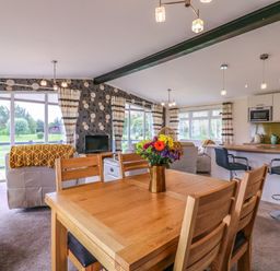 A dining area with wooden table and chairs adjacent to a living room with sofas and a TV and an open kitchen with bar stools at Owls Retreat Lodge 11 Near Aldeburgh