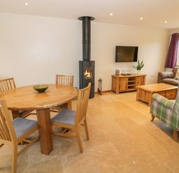 A dining table with four chairs next to a wood burning stove and a living area with a sofa a green armchair a coffee table and a TV at Lavender Cottage in Oxhill near Tysoe