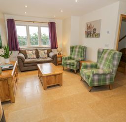 A living room with a brown sofa and two green plaid armchairs a wooden coffee table and side tables a wall mounted television and a wood burning stove at Lavender Cottage in Oxhill near Tysoe