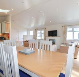 A dining table with white chairs in an open kitchen and living room with sofas and a wall-mounted TV at 43 The Maples in Sticker