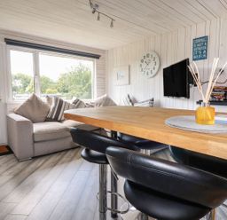 A living area with a gray sofa with cushions a wooden table with black chairs a wall clock a mounted television and a window with a view at Villa 18 Lanteglos Holiday Park in Lanteglos Holiday Park near Camelford