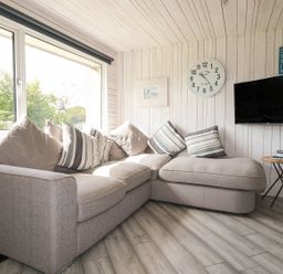 A living room with a sectional sofa and cushions a wall clock a TV and a side table with books at Villa 18 Lanteglos Holiday Park near Camelford