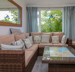 A living room with a brown wicker L shaped sofa with beige cushions and patterned pillows a glass coffee table striped curtains and a window with a fish decoration at 4 The Park in Kilkhampton