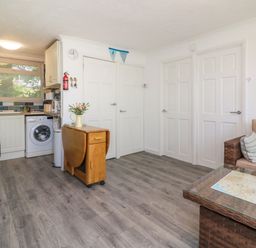 An open kitchen and living area with white doors a washing machine a small wooden foldable table and a wicker sofa with cushions at 4 The Park in Kilkhampton