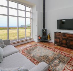 A living room with a large window, a gray sofa, a patterned rug, a wood stove, and a television on a wooden cabinet at West Dairy near Newport Pembrokeshire